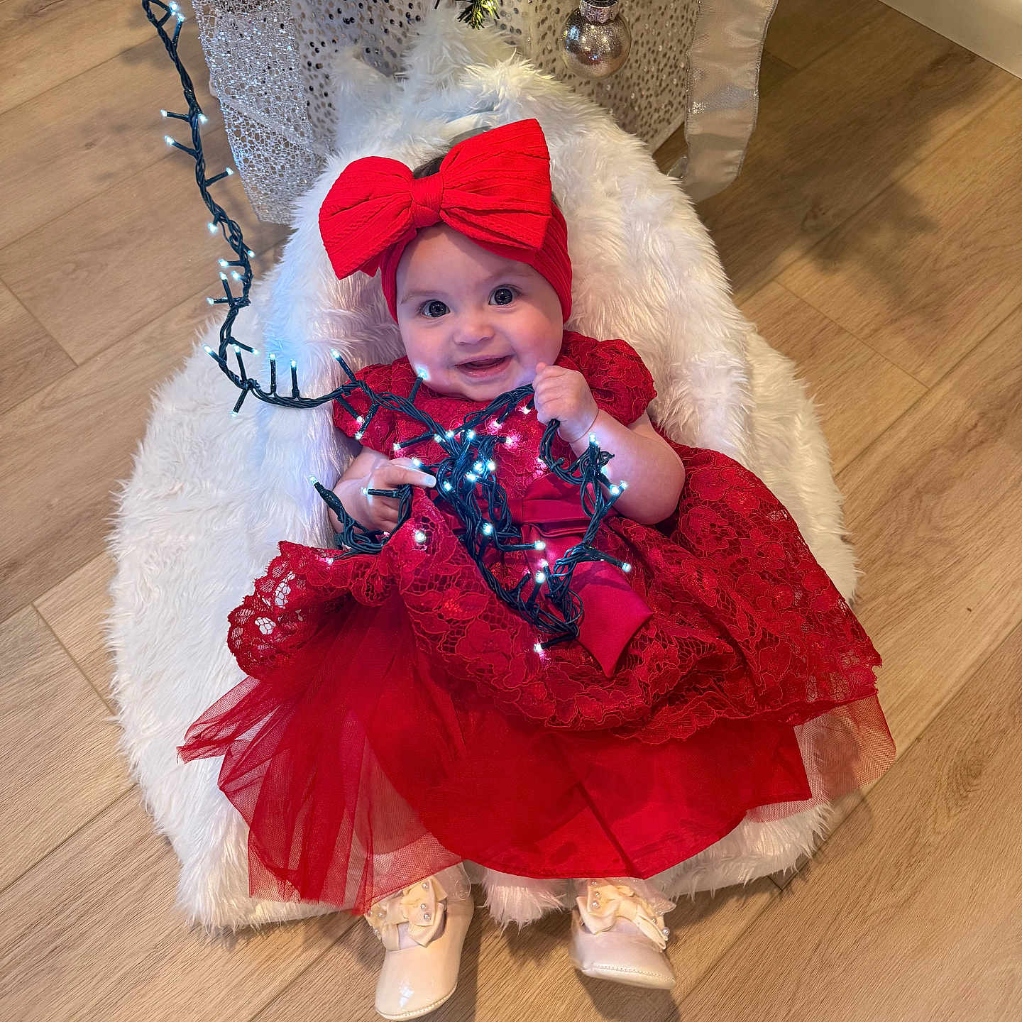 Cora joined the competition — help win amazing prizes! baby, child, red_dress, headband, bow, christmas_lights, holiday, smiling, indoor, flooring, wooden_floor, decorations, christmas_tree, festive, cushion, white_fur, cute, happy, portrait, toddler