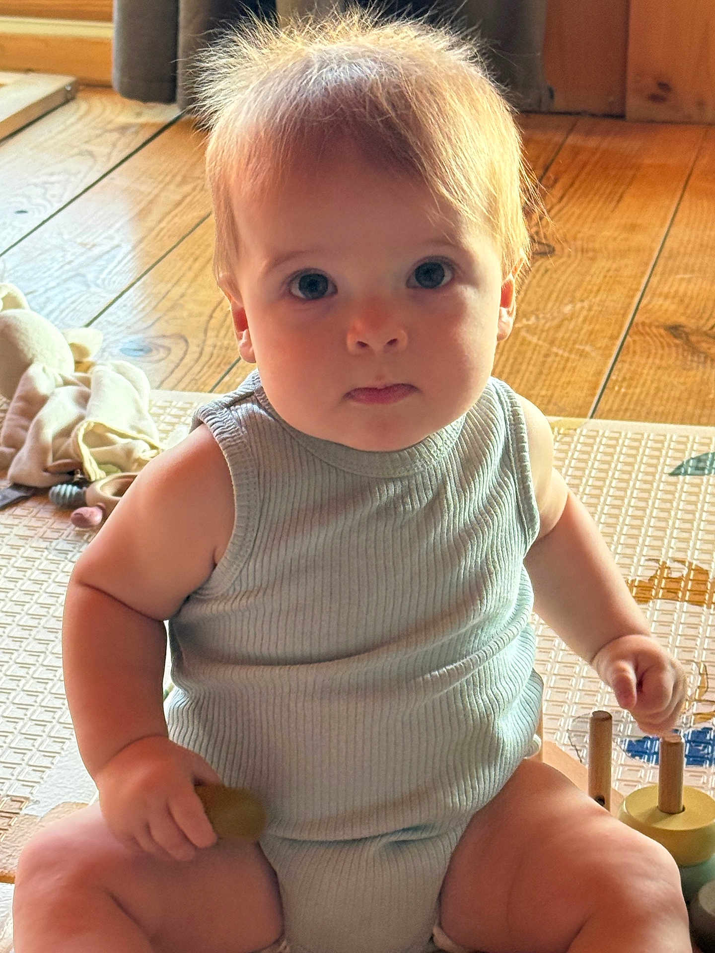 Timéo a rejoint le concours — aidez-le/la à gagner de superbes lots ! baby, child, infant, sitting, toy, play_mat, wooden_floor, natural_light, indoors, cute, chubby, face, hand, clothing, tank_top, expressionless, curious, hair, person, skin