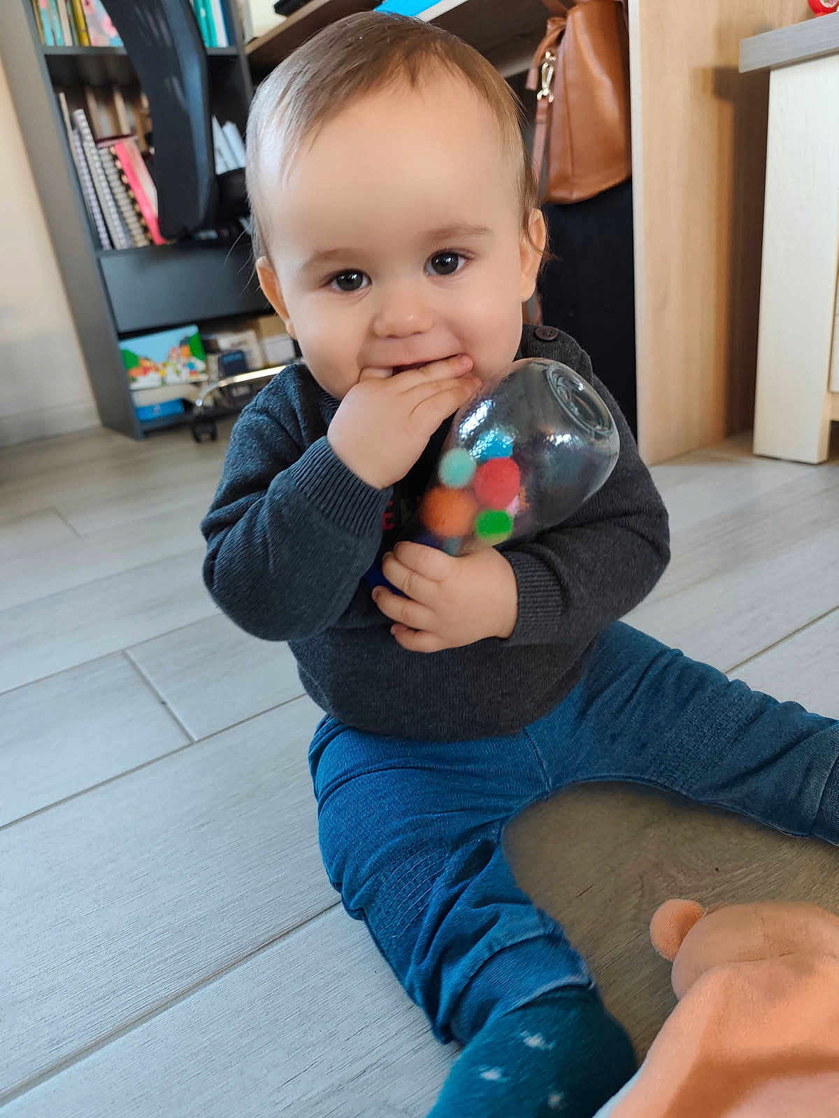 Kaïs a rejoint le concours — aidez-le/la à gagner de superbes lots ! baby, child, indoor, floor, toy, bottle, pom_poms, sweater, pants, sitting, hand, face, fingers, furniture, bookshelf, bag, playful, cute, person, smiling
