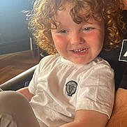 Ojay is registered to the contest to win money with this photo: child, toddler, curly_hair, smiling, white_shirt, indoor, natural_light, happy, person, face, rosy_cheeks, sitting, casual_clothing, logo, closeup, portrait, cozy, warm_light, cute, young