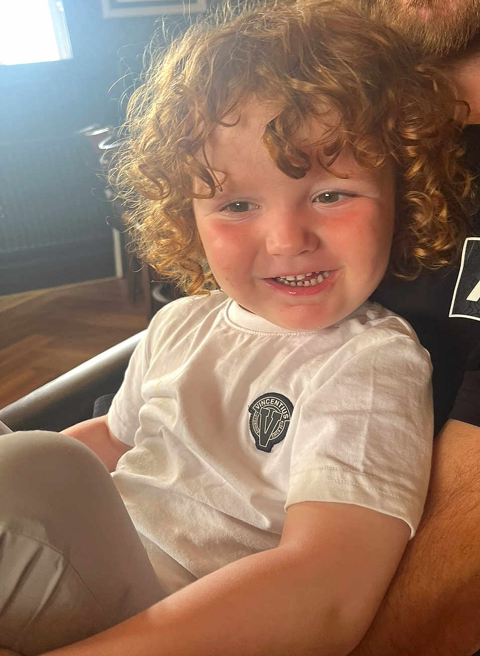 Ojay is registered to the contest to win money with this photo: child, toddler, curly_hair, smiling, white_shirt, indoor, natural_light, happy, person, face, rosy_cheeks, sitting, casual_clothing, logo, closeup, portrait, cozy, warm_light, cute, young