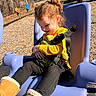 boots, casual, child, curly_hair, daylight, happy, nature, outdoor, park, play_equipment, playground, seatbelt, smiling, stuffed_animal, swing, toy, trees, winter_clothing, yellow_clothing, young_child