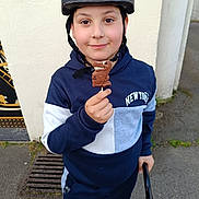 Nacim a rejoint le concours — aidez-le/la à gagner de superbes lots ! child, boy, helmet, chocolate, lollipop, outdoor, pavement, wall, smile, hoodie, blue_clothing, toy, sweet, person, young, standing, casual, happy, hand, helmet_strap