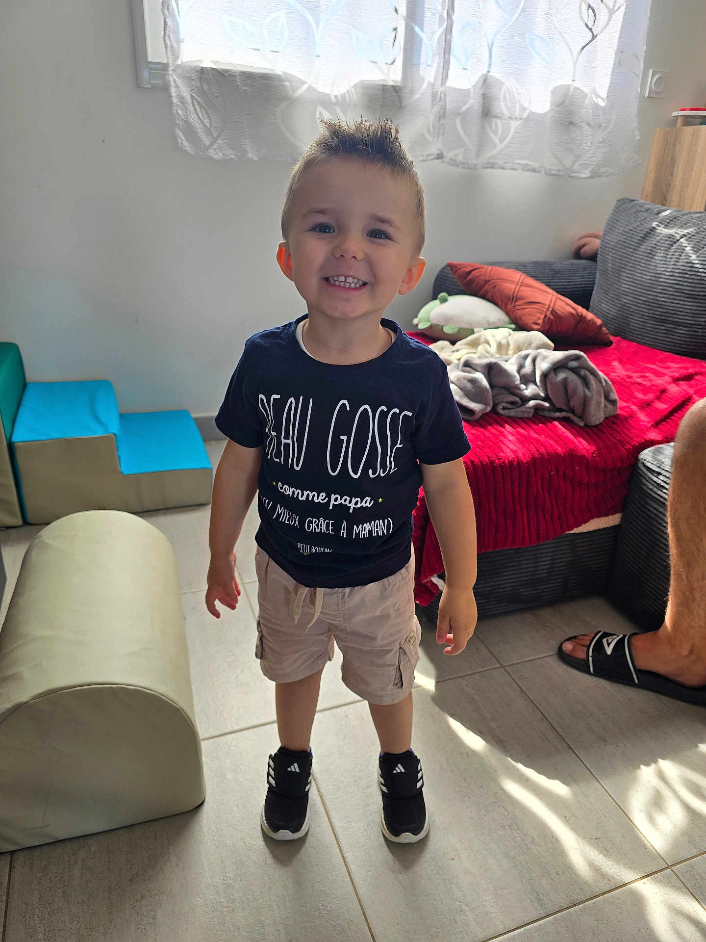 Eden a rejoint le concours — aidez-le/la à gagner de superbes lots ! child, toddler, smiling, indoor, casual_clothing, shorts, sneakers, curtains, couch, blanket, floor, window, toy, footwear, person, happy, hairstyle, living_room, home, sunlight