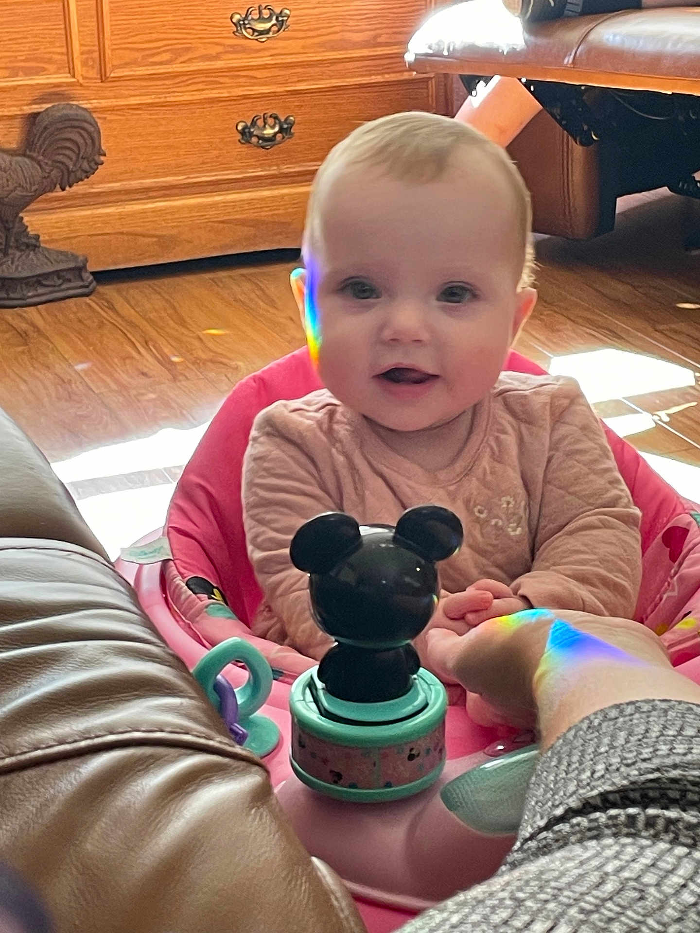 Luci is registered to the contest to win money with this photo: baby, child, pink_baby_seat, toy, mickey_mouse, rainbow_reflection, hand, wooden_floor, wooden_drawer, leather_sofa, indoor, happy, smiling, infant, person, cute, playful, holding_hand, home, cozy