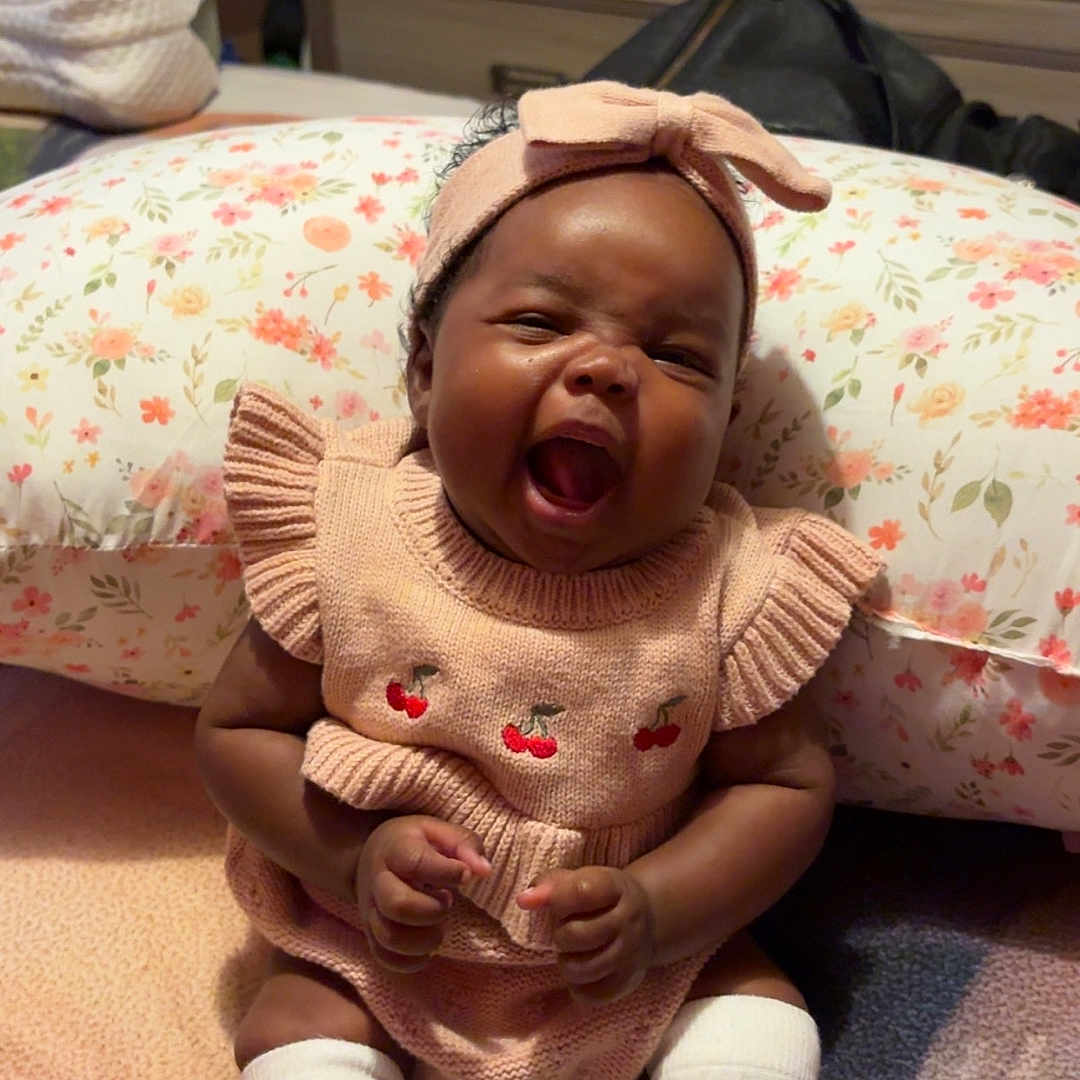 Kamoria is registered to the contest to win money with this photo: baby, child, infant, pink_outfit, headband, knitted_clothing, white_socks, floral_pillow, blanket, yawning, happy, indoors, furniture, dresser, person, cute, smiling, seated, cozy, home