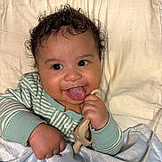 Shiloh joined the competition — help win amazing prizes! baby, curly_hair, tongue_out, toy, blanket, striped_clothing, bedspread, hand, smile, infant, cute, happy, indoors, closeup, face, child, soft_texture, lying_down, cozy, playful