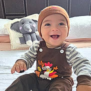 Emiliano is registered to the contest to win money with this photo: baby, child, smile, beanie, striped_clothing, plush_toy, elephant_toy, bed, pillow, knit_hat, happy, face, infant, sitting, indoor, cute, young_child, toy, button, cozy