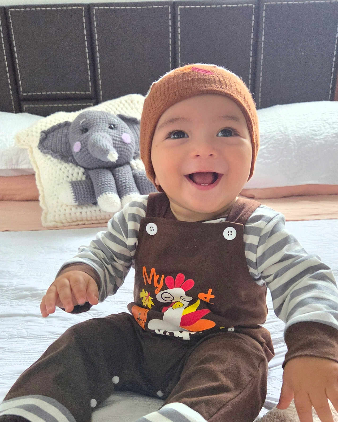 Emiliano is registered to the contest to win money with this photo: baby, child, smile, beanie, striped_clothing, plush_toy, elephant_toy, bed, pillow, knit_hat, happy, face, infant, sitting, indoor, cute, young_child, toy, button, cozy