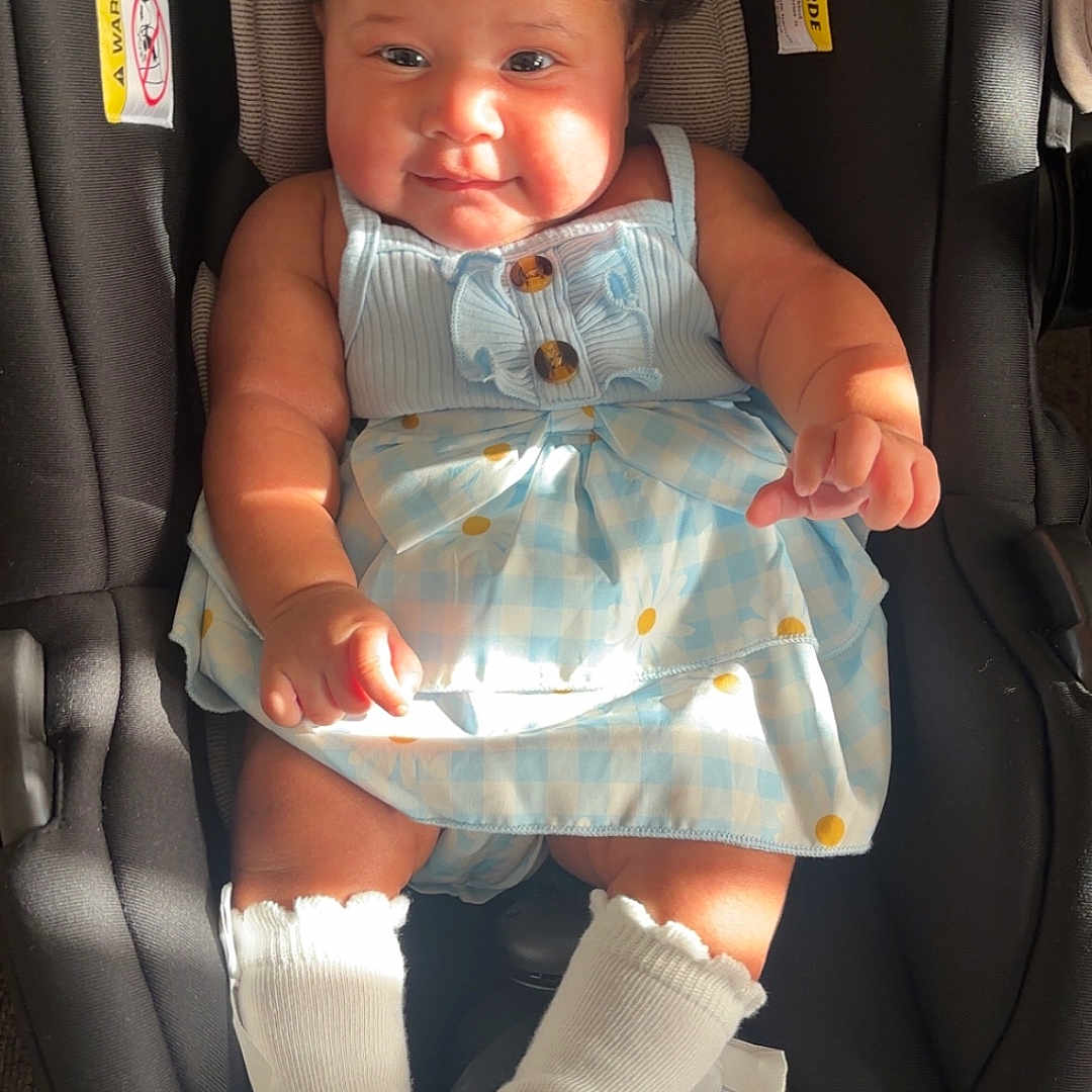 Emily is registered to the contest to win money with this photo: baby, infant, child, portrait, car_seat, sitting, dress, blue_dress, white_bows, hair_bows, socks, smiling, facial_expression, indoor, sunlight, vehicle, safety_seat, fashion, wood_floor, photograph
