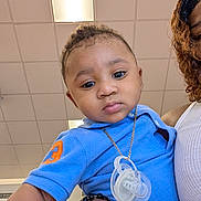 Chozen is registered to the contest to win money with this photo: adult, baby, blue_shirt, casual, ceiling, child, close_up, curly_hair, expression, face, fluorescent_light, gold_chain, hand, holding, indoor, pacifier, person, portrait, skin, white_top