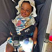 Louie is registered to the contest to win money with this photo: toddler, child, smiling, stroller, hat, sunhat, outdoor, baby, seatbelt, clothing, cute, happy, person, infant, daylight, sitting, young_child, baby_toys, colorful, portrait