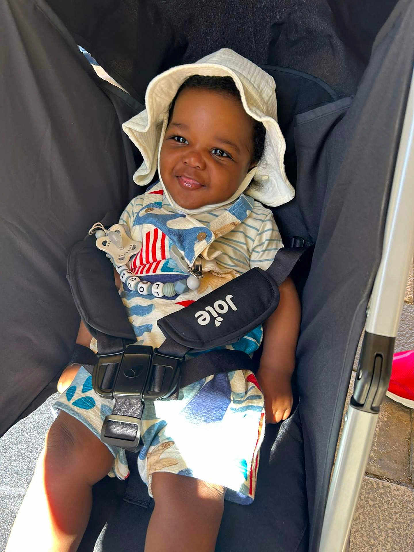 Louie is registered to the contest to win money with this photo: toddler, child, smiling, stroller, hat, sunhat, outdoor, baby, seatbelt, clothing, cute, happy, person, infant, daylight, sitting, young_child, baby_toys, colorful, portrait