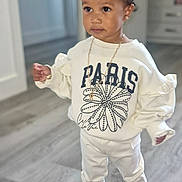 Renaya-Rose is registered to the contest to win money with this photo: toddler, child, indoors, standing, white_clothing, sweatshirt, pants, black_shoes, necklace, hairstyle, topknot, flooring, wood_floor, curious, casual_wear, young_child, person, portrait, home_interior, fashion