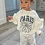 toddler, child, indoors, standing, white_clothing, sweatshirt, pants, black_shoes, necklace, hairstyle, topknot, flooring, wood_floor, curious, casual_wear, young_child, person, portrait, home_interior, fashion