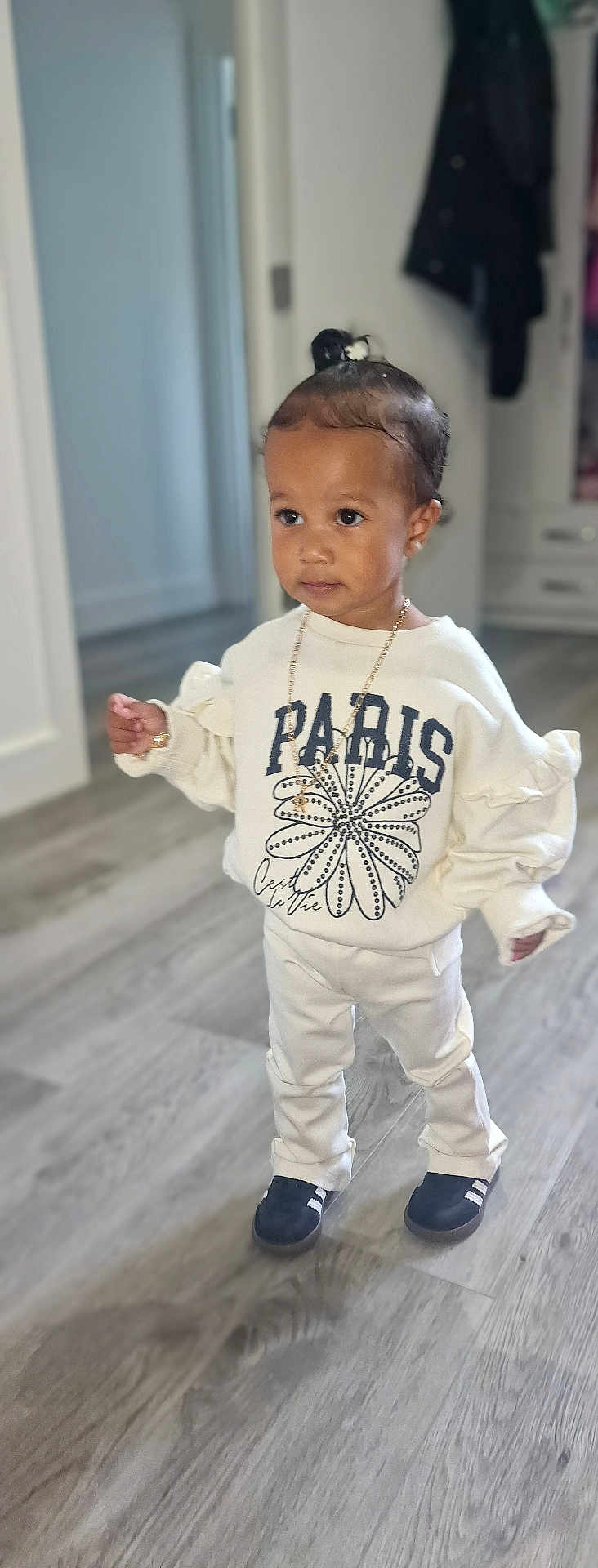 Renaya-Rose is registered to the contest to win money with this photo: toddler, child, indoors, standing, white_clothing, sweatshirt, pants, black_shoes, necklace, hairstyle, topknot, flooring, wood_floor, curious, casual_wear, young_child, person, portrait, home_interior, fashion