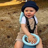 Lyam participe au concours pour gagner de l'argent avec cette photo : bucket_hat, casual_clothing, child, dirt, face, hand, happy, hat, nature, outdoor, overalls, park, person, pine_needles, play, ride_on_toy, seat, smile, sunlight, toddler