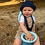 bucket_hat, casual_clothing, child, dirt, face, hand, happy, hat, nature, outdoor, overalls, park, person, pine_needles, play, ride_on_toy, seat, smile, sunlight, toddler