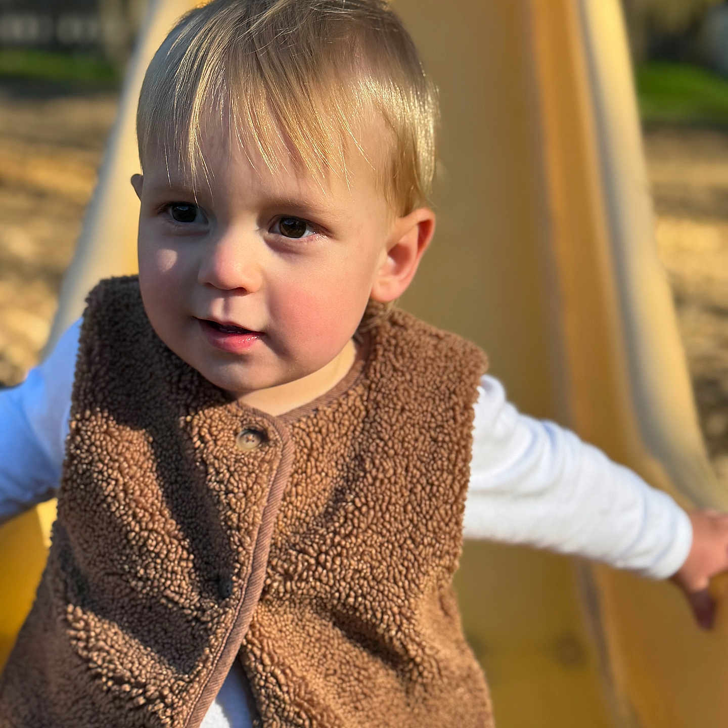 Louis participe au concours pour gagner de l'argent avec cette photo : child, toddler, blonde_hair, brown_vest, white_shirt, slide, playground, outdoor, sunlight, face, portrait, curious_expression, rosy_cheeks, daylight, playing, clothing, smiling, skin, person, fun