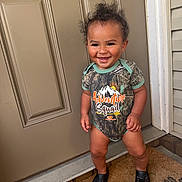 Koa is registered to the contest to win money with this photo: toddler, child, smile, standing, curly_hair, onesie, camouflage, shoes, doormat, door, porch, happy, outdoor, person, cute, expression, face, feet, clothing, home