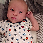 Carter is registered to the contest to win money with this photo: baby, infant, face, eyes, clothing, onesie, pattern, sports_balls, blanket, fabric, texture, person, child, cute, portrait, lying_down, indoor, soft, skin, newborn