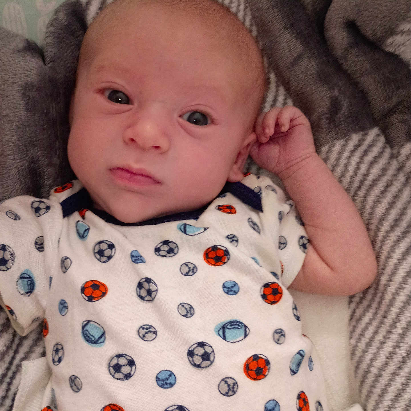 Carter is registered to the contest to win money with this photo: baby, infant, face, eyes, clothing, onesie, pattern, sports_balls, blanket, fabric, texture, person, child, cute, portrait, lying_down, indoor, soft, skin, newborn