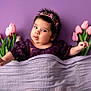 adorable, baby, blanket, child, chubby_cheeks, closeup, clothing, curious_expression, cute, floral, flower_crown, headband, indoor, infant, laying_down, lilac, pink_tulips, portrait, purple_background, soft_texture