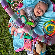 Khloe is registered to the contest to win money with this photo: baby, smiling, blue_blanket, outdoor, grass, toys, headband, baby_playmat, colorful, infant, happy, cute, stuffed_animal, child, playtime, person, nature, baby_clothes, soft_toys, relaxing
