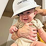 adult, apparel, baby, child, closeup, construction_helmet, cute, drool, face, hands, happy, headwear, helmet, holding, indoor, infant, onesie, person, skin, smiling
