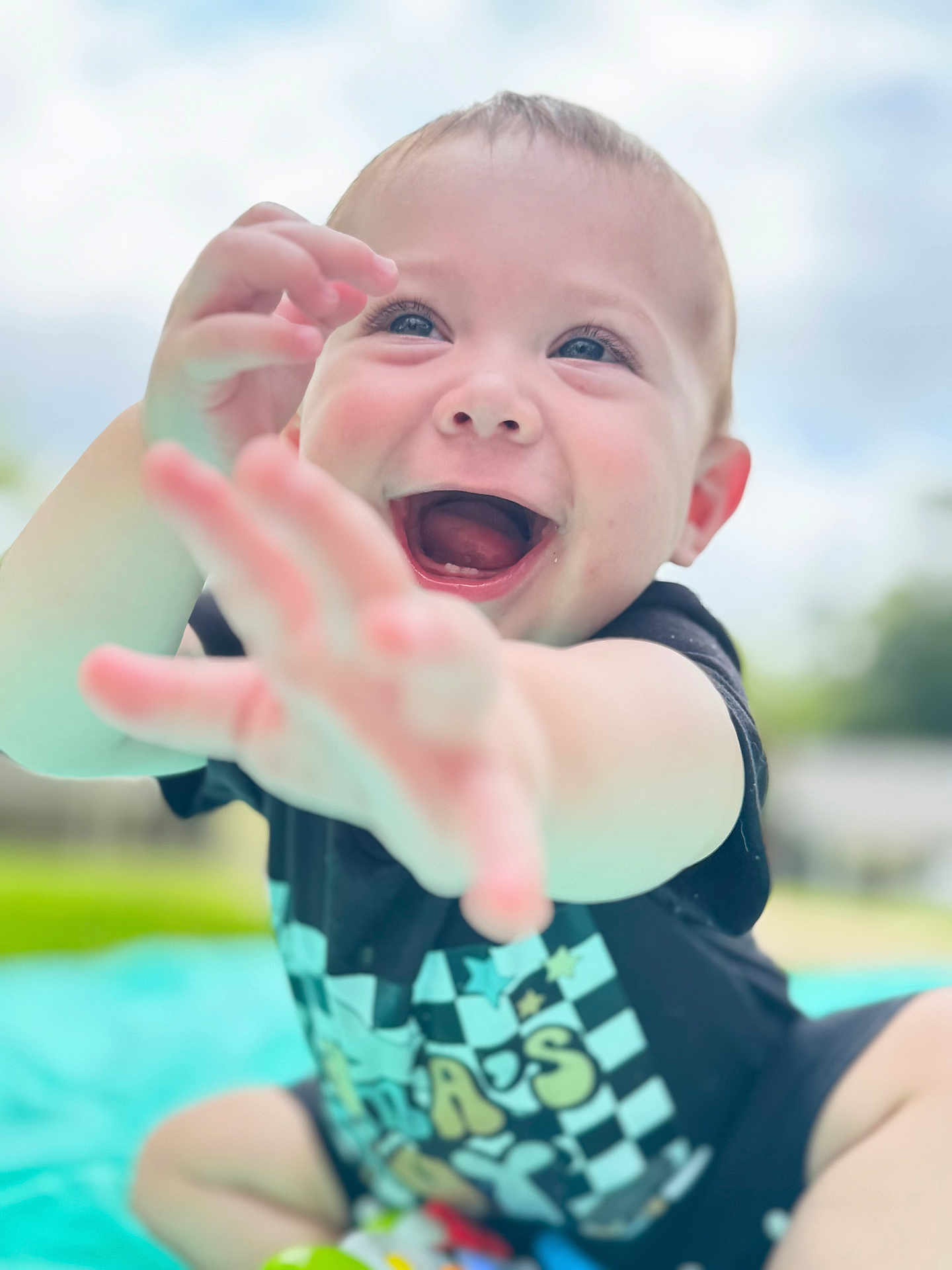 Waylon joined the competition — help win amazing prizes! baby, child, smiling, happy, reaching, outdoor, sky, greenery, shirt, blue_blanket, portrait, cute, young, joyful, expressive, sitting, face, hands, daylight, blurred_background