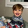 child, toddler, smiling, sitting, ball_pit, balls, pastel_colors, sweater, festive_clothing, indoor, window, happy, face, person, playing, cozy, cute, joyful, portrait, holiday