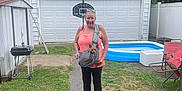 Cookie is registered to the contest to win money with this photo: woman, dog, backyard, garage, basketball_hoop, inflatable_pool, grass, pathway, trees, summer, casual_clothing, flip_flops, pet_carrier, smiling, outdoor, daylight, recreation, relaxation, small_dog, leisure