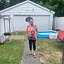 woman, dog, backyard, garage, basketball_hoop, inflatable_pool, grass, pathway, trees, summer, casual_clothing, flip_flops, pet_carrier, smiling, outdoor, daylight, recreation, relaxation, small_dog, leisure