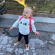 Camila a rejoint le concours — aidez-le/la à gagner de superbes lots ! toddler, child, outdoor, pavement, backpack, sweater, pigtails, shoes, curly_hair, sunlight, shadow, fence, concrete, wall, playful, standing, happy, casual_clothing, young_child, smiling