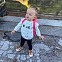 toddler, child, outdoor, pavement, backpack, sweater, pigtails, shoes, curly_hair, sunlight, shadow, fence, concrete, wall, playful, standing, happy, casual_clothing, young_child, smiling