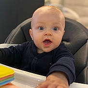 Tilio a rejoint le concours — aidez-le/la à gagner de superbes lots ! baby, child, infant, high_chair, reaching_hand, blue_eyes, black_clothing, indoor, toy, colorful, face, person, cute, expression, young_child, skin, head, mouth_open, seated, focus
