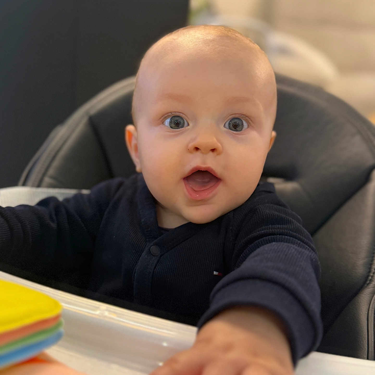 Tilio a rejoint le concours — aidez-le/la à gagner de superbes lots ! baby, black_clothing, blue_eyes, child, colorful, cute, expression, face, focus, head, high_chair, indoor, infant, mouth_open, person, reaching_hand, seated, skin, toy, young_child