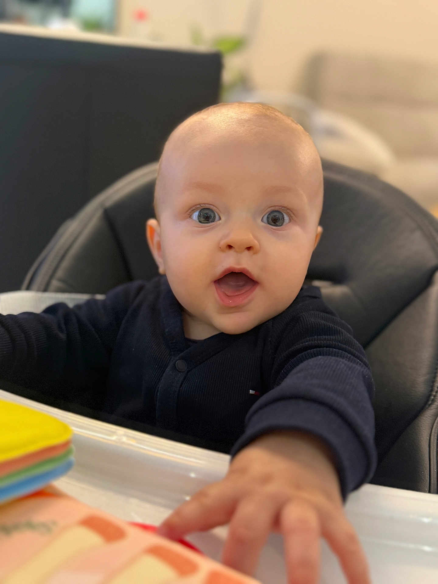 Tilio a rejoint le concours — aidez-le/la à gagner de superbes lots ! baby, child, infant, high_chair, reaching_hand, blue_eyes, black_clothing, indoor, toy, colorful, face, person, cute, expression, young_child, skin, head, mouth_open, seated, focus