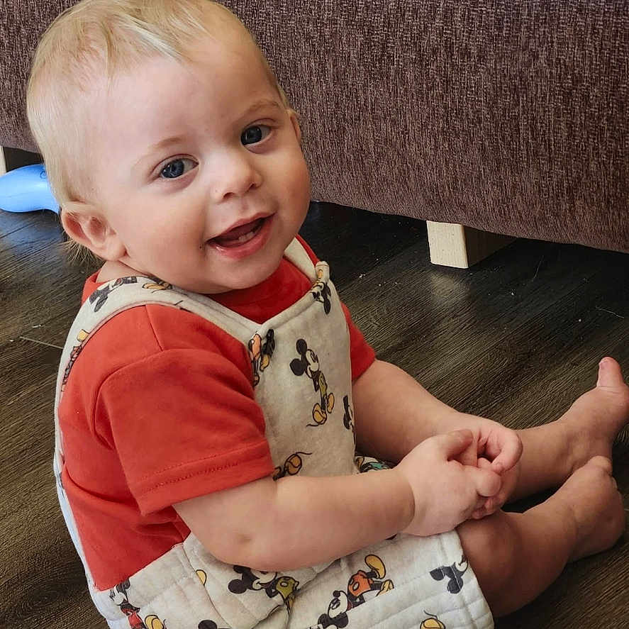 Jayce is registered to the contest to win money with this photo: toddler, child, smiling, sitting, floor, overalls, mickey_mouse, red_shirt, blond_hair, blue_eyes, indoor, wooden_floor, couch, furniture, cute, happy, baby, portrait, casual_clothing, playful