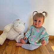 Enora a rejoint le concours — aidez-le/la à gagner de superbes lots ! toddler, child, pigtails, smile, blue_eyes, stuffed_animal, plush_hippo, wooden_floor, indoor, sweater, floral_pattern, leggings, barefeet, sitting, portrait, sunlight, shadow, wall, cozy, cute