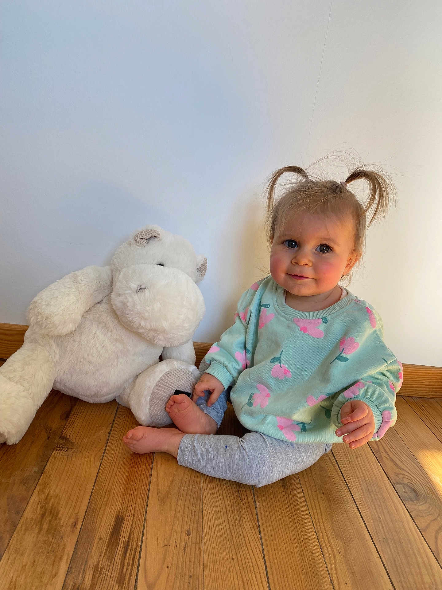 Enora a rejoint le concours — aidez-le/la à gagner de superbes lots ! toddler, child, pigtails, smile, blue_eyes, stuffed_animal, plush_hippo, wooden_floor, indoor, sweater, floral_pattern, leggings, barefeet, sitting, portrait, sunlight, shadow, wall, cozy, cute