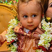Florentin participe au concours pour gagner de l'argent avec cette photo : toddler, child, flower_garland, traditional_clothing, embroidery, gold_chain, person, hand, ring, face, curly_hair, blue_eyes, indoor, colorful_background, portrait, closeup, celebration, decor, seated, cultural