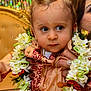 toddler, child, flower_garland, traditional_clothing, embroidery, gold_chain, person, hand, ring, face, curly_hair, blue_eyes, indoor, colorful_background, portrait, closeup, celebration, decor, seated, cultural