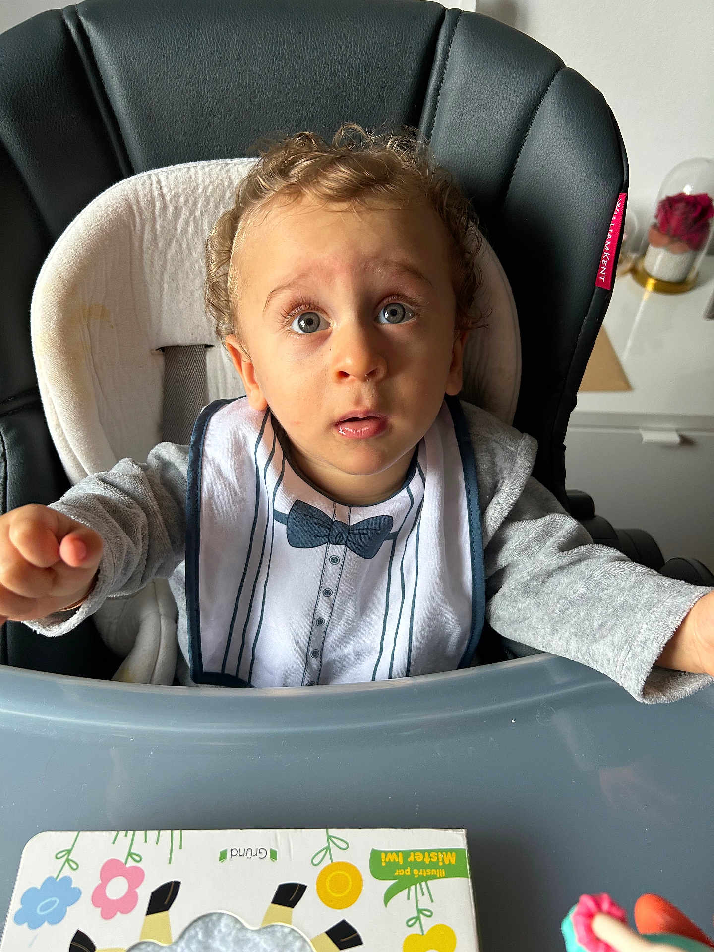 Florentin participe au concours pour gagner de l'argent avec cette photo : baby, toddler, high_chair, bib, curious, indoor, toy, table, face, child, clothing, expression, seat, curly_hair, person, furniture, tray, pink, blue, gray