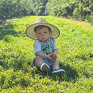 Cody is registered to the contest to win money with this photo: toddler, child, hat, apple, grass, outdoor, nature, greenery, sunlight, orchard, baby, clothing, footwear, portrait, sitting, summer, daytime, cute, young_child, casual