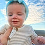 Millie is registered to the contest to win money with this photo: baby, child, headband, blue, striped_clothing, smile, face, hand, outdoor, sky, clouds, daylight, cute, infant, person, portrait, buttoned_clothing, closeup, happy, young