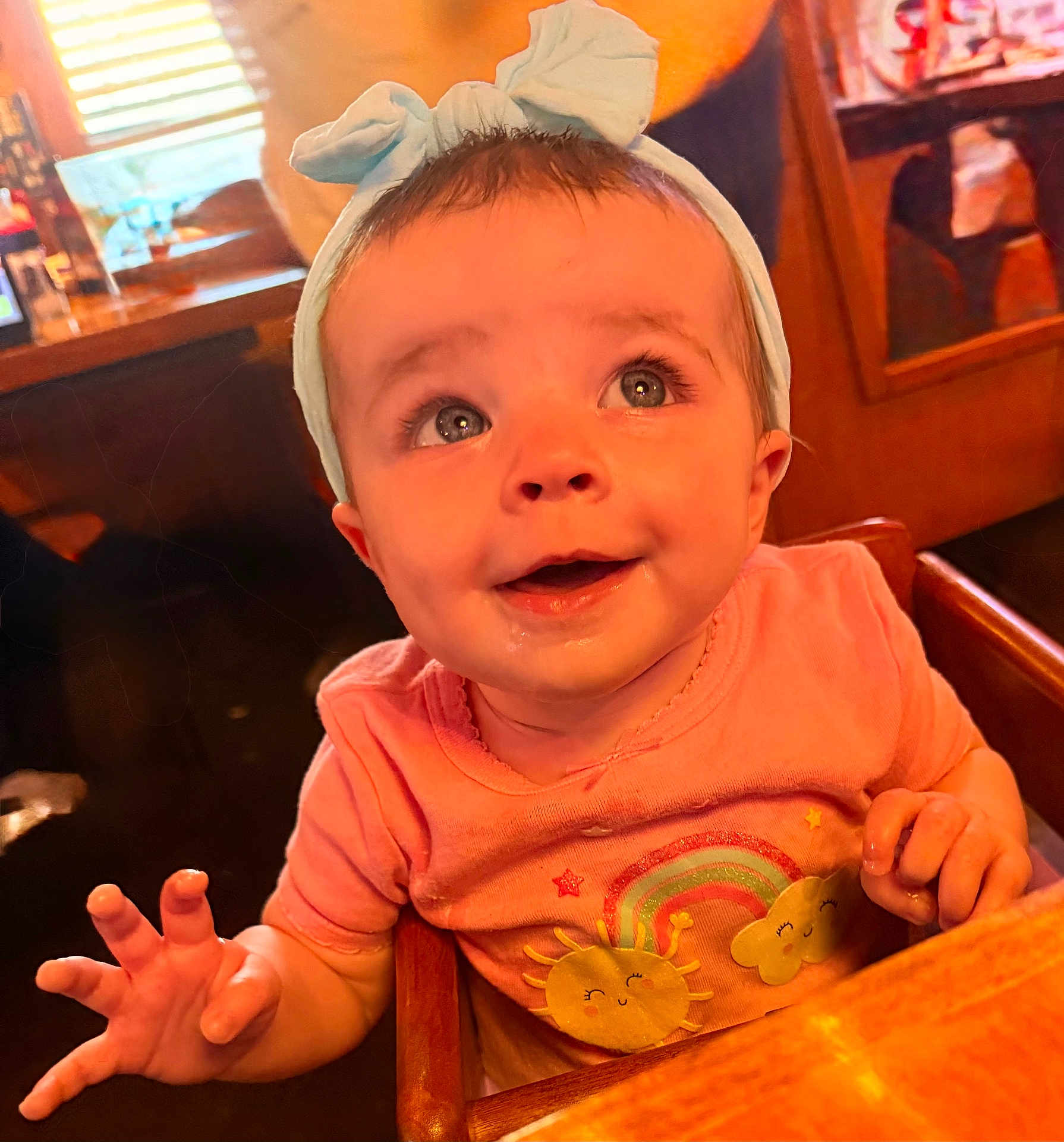 Millie is registered to the contest to win money with this photo: baby, child, infant, headband, bow, pink_shirt, sun, cloud, rainbow, high_chair, wooden_furniture, indoor, cute, smiling, happy, portrait, closeup, person, expression, reaching_hand