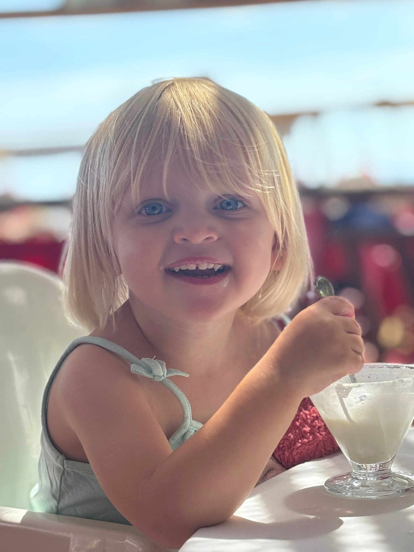 Julia participe au concours pour gagner de l'argent avec cette photo : child, toddler, blonde_hair, blue_eyes, smile, spoon, dessert, glass_cup, ice_cream, high_chair, dining_table, close_up, portrait, candid, happy, bangs, sleeveless_top, soft_lighting, background_bokeh, family_restaurant