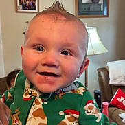 David joined the competition — help win amazing prizes! baby, child, smiling, happy, face, person, green_clothing, festive, reindeer_pattern, santa_hat, onesie, indoor, living_room, lamp, hand, portrait, cute, holiday, moawk_hairstyle, cozy