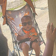 Cadence is registered to the contest to win money with this photo: baby, sunglasses, stroller, child, hand, outdoor, sunlight, wooden_post, footwear, baby_bib, moose_pattern, trees_pattern, curious, candid, summer, shade, infant, toddler, grip, relaxation